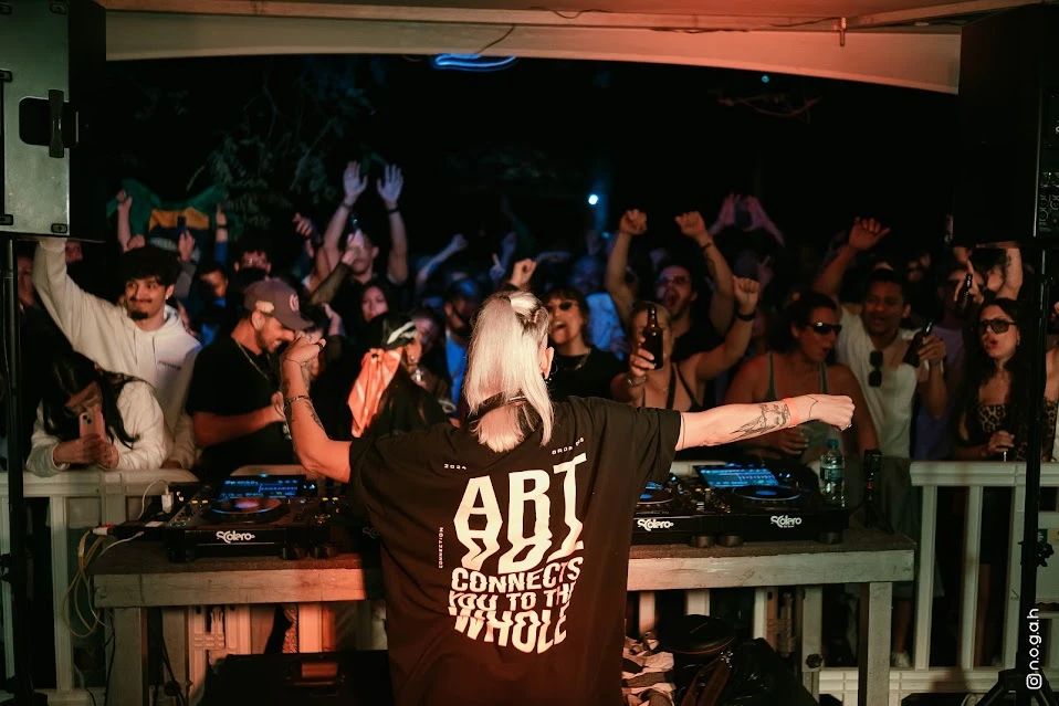 DJ de costas, com a camiseta “Art connects you to the whole”, e o público de braços para cima
