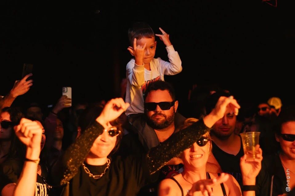 Criança nos ombros do pai no meio do público, fazendo sinal de rock com as mãos