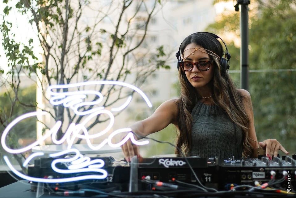 DJ tocando ao lado do logo da Cura em neon, num fim de tarde