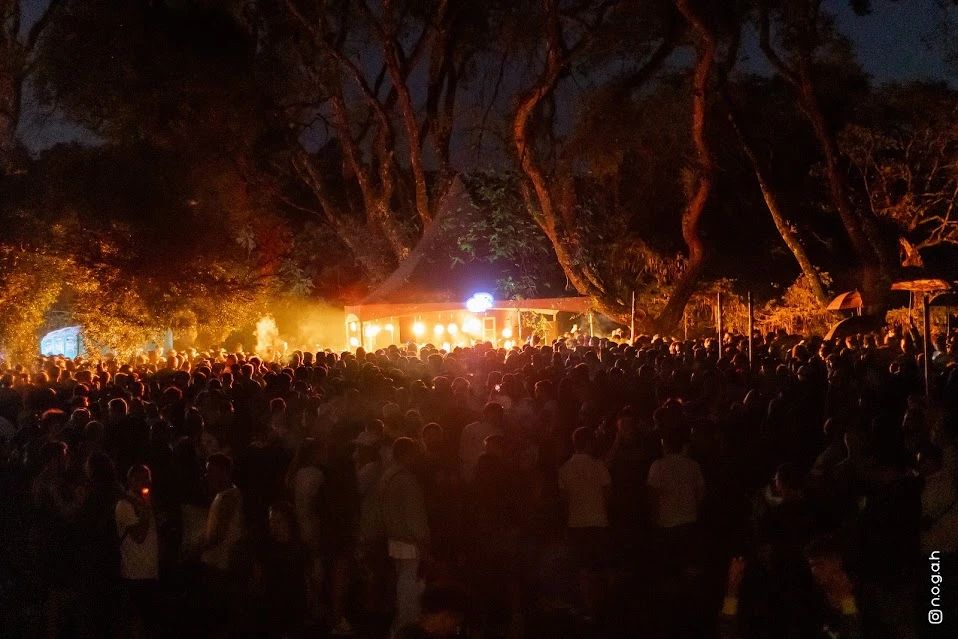 A pista lotada no parque depois do pôr do sol, com a tenda iluminada entre as árvores